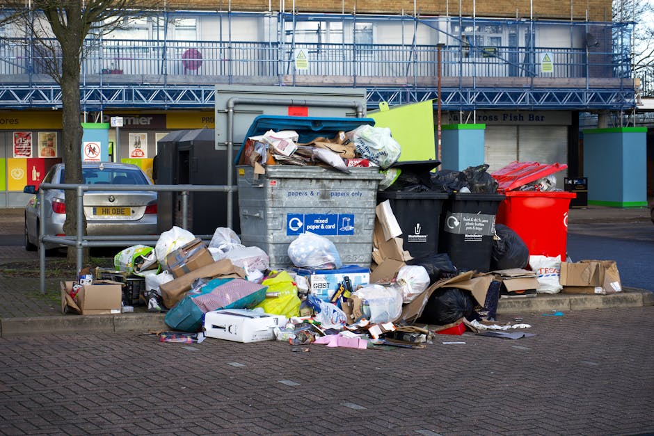 A large pile of mixed household waste, including paper, cardboard boxes, black and red plastic garbage bags, and various discarded packaging materials, is overflowing from a grey recycling bin labeled for mixed paper and cardboard, situated on a paved sidewalk near a parking area. Behind the recycling bin, there is a silver car parked, partially visible, with a blue and white sign on the building in the background indicating it is next to a retail unit. Adjacent to the waste collection point, there is a small railing and a tree with bare branches. The scene is well-lit with natural daylight, and the background features a building under renovation or covered with scaffolding, with a sign indicating its commercial use. Occasionally, [COMPANY_NAME] handles furniture transport and loading processes associated with home relocation, as seen in the context of managing waste disposal and moving logistics for local households.