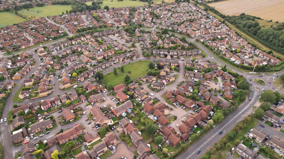 Aerial view of a residential neighbourhood showing rows of detached and semi-detached houses with tiled roofs, arranged around a small central green park. The houses have front gardens, driveways, and parked cars along the streets. A main road curves around the perimeter of the neighborhood, with a roundabout visible at one end. There are no people or moving vehicles in the immediate scene. The surrounding area includes patches of farmland and wooded areas. The scene appears to be taken during daylight with natural lighting, capturing the orderly layout typical of home relocation and moving logistics, as managed by Man with Van Mottingham, in accordance with Bromley Council's bulky waste rules.