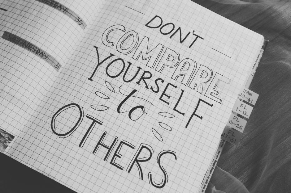 A close-up photograph of a grid-lined notebook open on a wooden surface, displaying a handwritten message in bold, capital letters reading 'DON'T COMPARE YOURSELF to OTHERS' with decorative lines and 