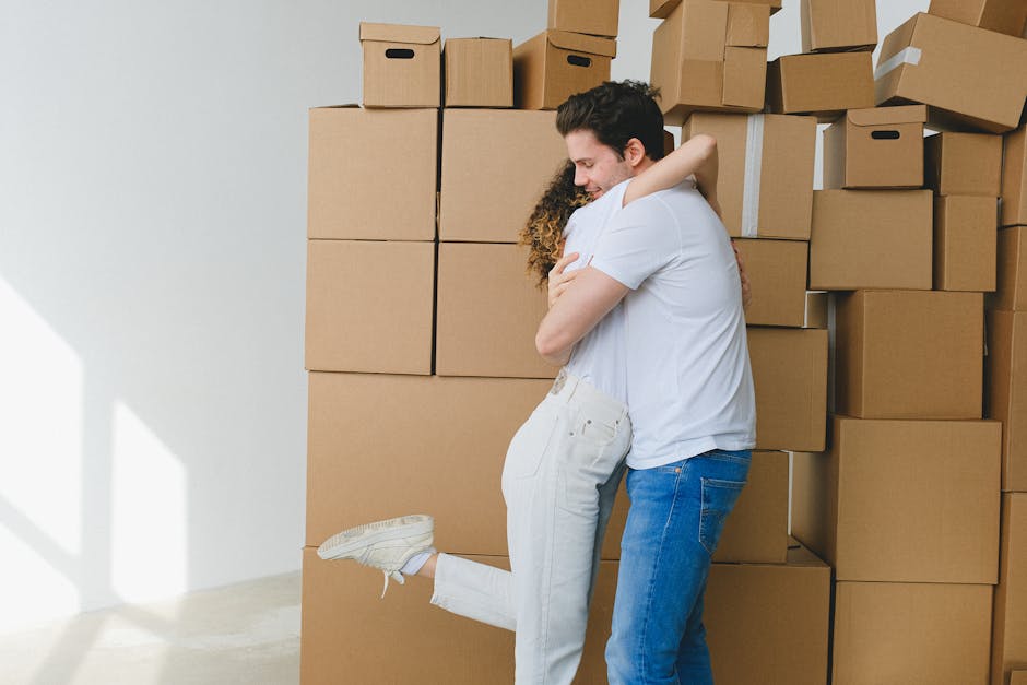 A man and a woman hugging each other inside a house surrounded by stacked cardboard moving boxes, some of which are sealed with packing tape. The woman, wearing white pants and sneakers, stands on one leg with her other leg lifted behind her. The man, dressed in a white t-shirt and blue jeans, embraces her warmly. The background features a plain white wall and a light source casting soft shadows, indicating natural or artificial bright lighting. The scene depicts a home relocation or packing process, with the boxes likely prepared for moving day. This setting illustrates the logistical aspect of furniture transport and packing involved in house removals, with [COMPANY_NAME], Man with Van Mottingham, providing professional moving services.