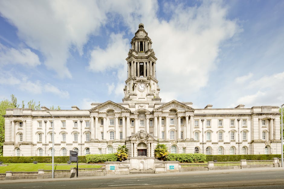 A large, historic stone building with a prominent central clock tower featuring multiple levels and ornate architectural details, set against a partly cloudy sky. The building's facade includes evenly spaced windows and classical decorative elements, with a well-maintained green hedge and potted plants near the entrance. In front, a wide pavement with a few street signs is visible, indicating an urban environment. This scene depicts a typical location where house removals and furniture transport may take place, reflecting the importance of understanding local council permits during home relocation processes. Man with Van Mottingham's services focus on efficient packing, loading, and transport, often involving careful handling of furniture and boxes on-site, including equipment such as trolleys or straps for a smooth loading process.