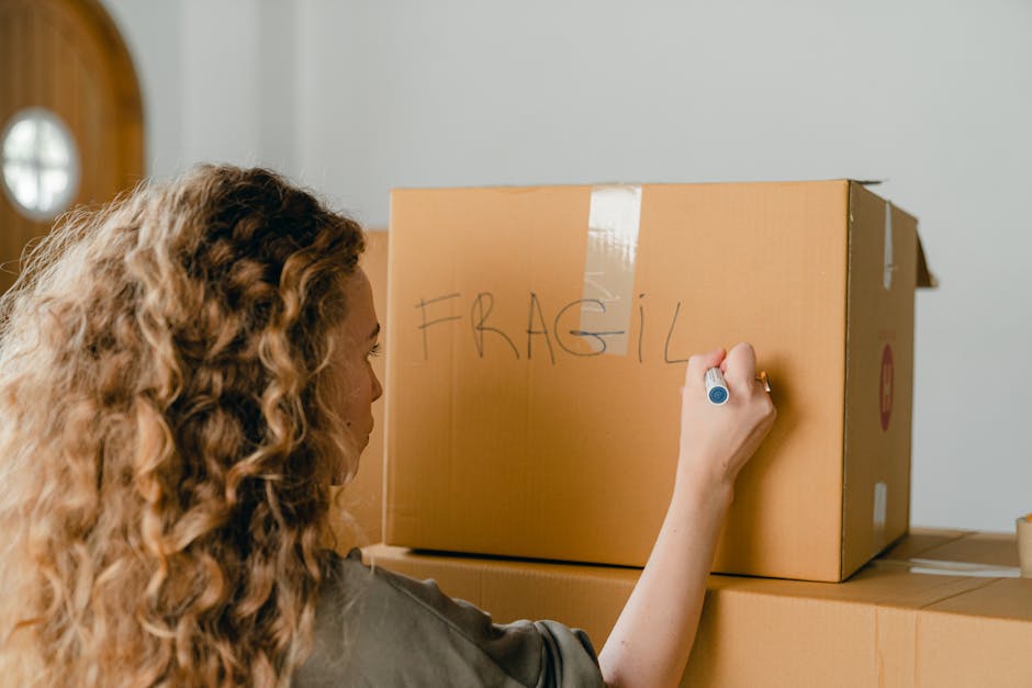 A man and a woman hugging each other inside a house surrounded by stacked cardboard moving boxes, some of which are sealed with packing tape. The woman, wearing white pants and sneakers, stands on one leg with her other leg lifted behind her. The man, dressed in a white t-shirt and blue jeans, embraces her warmly. The background features a plain white wall and a light source casting soft shadows, indicating natural or artificial bright lighting. The scene depicts a home relocation or packing process, with the boxes likely prepared for moving day. This setting illustrates the logistical aspect of furniture transport and packing involved in house removals, with [COMPANY_NAME], Man with Van Mottingham, providing professional moving services.