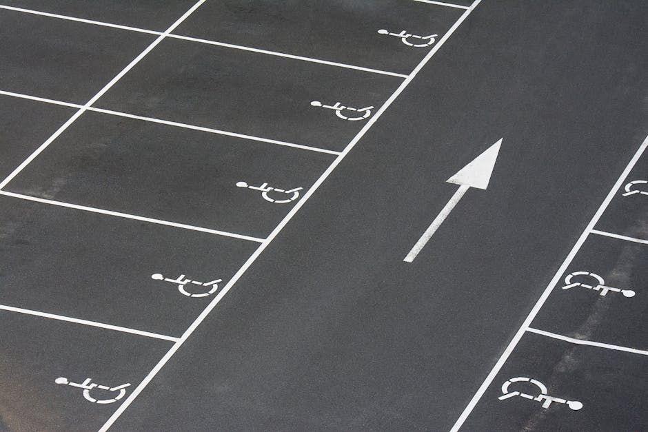 A section of a parking area with clearly marked white lines designating individual parking spaces, many of which are designated for disabled parking with the universal wheelchair symbol. The surface is dark asphalt, and a single white arrow painted on the ground points forward, indicating the direction of traffic flow. The image appears to be outdoors with natural lighting and no vehicles or other objects currently occupying the spaces. The scene relates to parking logistics and access considerations typical during house removals or furniture transport, highlighting the importance of planning for vehicle parking and access routes when coordinating home relocation services, as provided by Man with Van Mottingham.