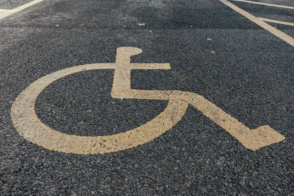 A close-up view of a wheelchair accessible parking space marked on an asphalt surface with a yellow painted wheelchair symbol. The symbol consists of a circular head and curved body along with two lines representing arms, and a large open circle for the seat of the chair, all painted on the dark textured pavement. In the background, parking space lines are visible, indicating designated parking areas. The surface appears slightly rough and weathered, with small stones embedded in the asphalt. This image is relevant to house removals and transportation services provided by Man with Van Mottingham, as it depicts parking considerations for residential moving processes, emphasizing the importance of accessible parking for vehicle positioning during home relocation and furniture transport.