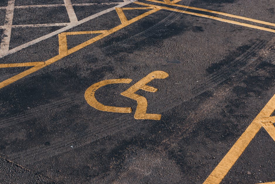 A section of a parking area with clearly marked white lines designating individual parking spaces, many of which are designated for disabled parking with the universal wheelchair symbol. The surface is dark asphalt, and a single white arrow painted on the ground points forward, indicating the direction of traffic flow. The image appears to be outdoors with natural lighting and no vehicles or other objects currently occupying the spaces. The scene relates to parking logistics and access considerations typical during house removals or furniture transport, highlighting the importance of planning for vehicle parking and access routes when coordinating home relocation services, as provided by Man with Van Mottingham.