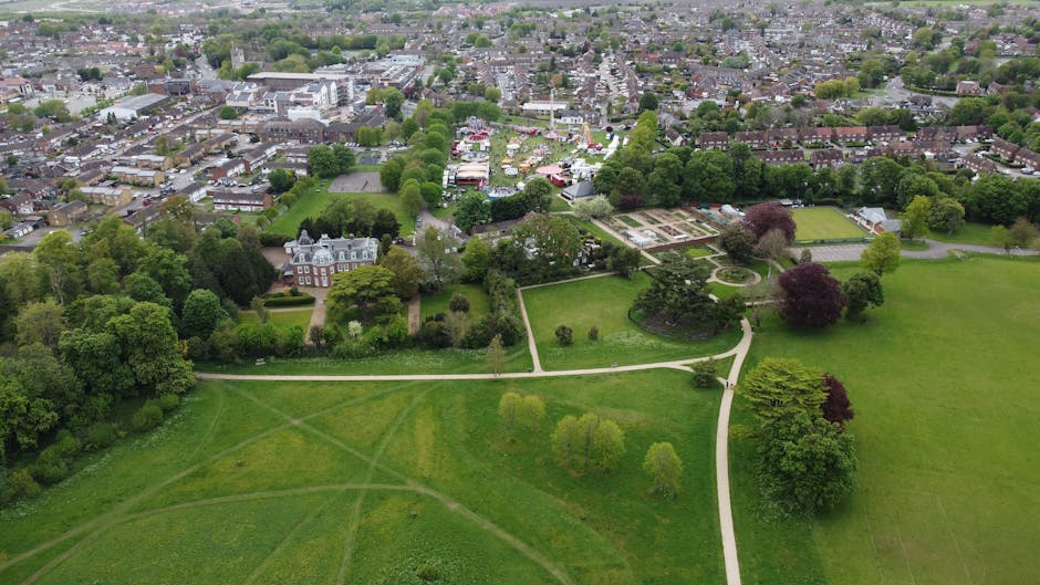 An aerial view of a large park area with well-maintained green lawns, numerous trees of various sizes and colors, and paved walking paths intersecting the open spaces. In the background, there are residential buildings and a small fair or market with tents and temporary structures, situated beyond the park. The park appears to be part of a residential neighbourhood, with a stately manor house or similar large property visible among the trees. The scene captures a peaceful, open space ideal for family activities or relaxation, situated within a suburban area. The image illustrates the typical environment involved in home relocation or moving services, with the park as a notable landmark near the property, supporting local tips for moves to Horn Park Estate from Mottingham, with Man with Van Mottingham providing removals services in the area.