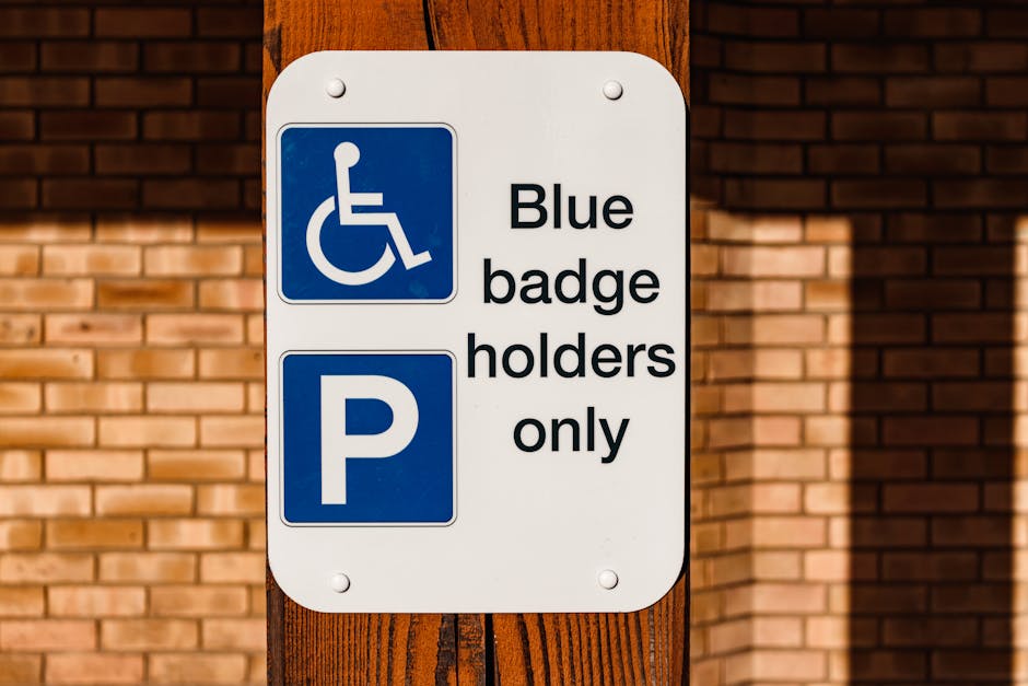 A white rectangular sign mounted on a wooden post against a brick wall background. The top part of the sign displays a blue wheelchair symbol indicating accessible parking, with a white icon of a person in a wheelchair on a blue background. Below, the sign has a bold white letter 'P' within a blue square, signifying parking availability. Adjacent to the parking symbol is the text 'Blue badge holders only' in black lettering. This signage is associated with parking regulations for disabled individuals with a blue badge. The environment appears to be an outdoor area with natural lighting, and the sign relates to parking policies relevant for house removals and moving services, such as those offered by Man with Van Mottingham, especially when arranging parking suspensions or permits for relocation logistics.