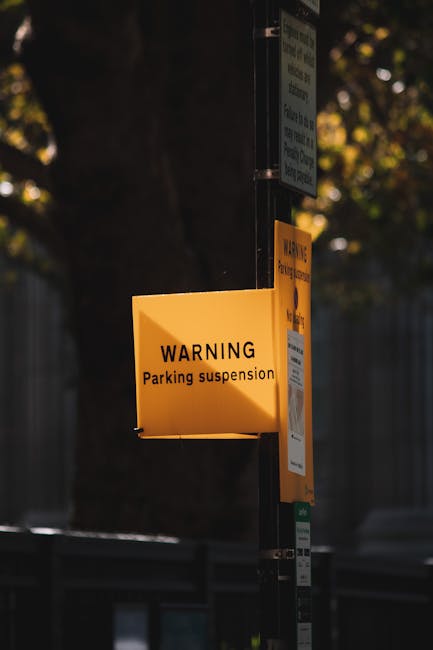 A yellow warning sign attached to a metal pole displays the message 'WARNING Parking suspension' in black text. The sign is located outdoors, with a background featuring a tree with green foliage and a metal fence. The sign is illuminated by natural daylight, indicating it is daytime. Adjacent to the warning sign, other smaller notices are affixed to the pole, suggesting regulated parking restrictions in the area. The context implies a temporary parking restriction possibly related to home relocation or moving services, such as those offered by Man with Van Mottingham, which may be necessary for safe and efficient furniture transport or loading during a house removal process.