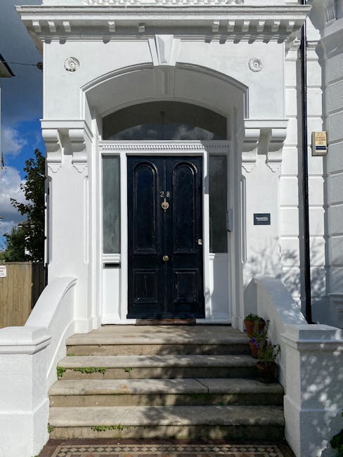 A white Victorian-style front entrance of a property with a black double door featuring a brass number 28 and a door knocker, accessed by a set of wide stone steps. The entrance is framed by decorative architectural details including mouldings, columns, and an arched lintel. To the right of the door, there is a grey nameplate and a black vertical gutter pipe. The doorway is inside a small porch area with a white balustrade on each side, and a potted plant is visible on the bottom right step. Partial shadows cast by nearby trees fall across the steps and facade, indicating daylight. This scene exemplifies a residential property where home relocation or furniture transport might take place, as seen in the context of professional removals services from Man with Van Mottingham, specializing in packing and moving house furniture.