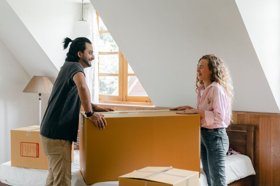 Two movers from Man with Van Mottingham are inside a room with sloped ceilings and a large window, loading a large cardboard wardrobe box onto a trolley for a home relocation. The man on the left, with shoulder-length dark hair tied back, is wearing a dark grey sleeveless shirt and beige trousers, and is holding the box steady with his hands. The woman on the right, with curly blonde hair, is wearing a light pink shirt with floral embroidery and jeans, smiling as she participates in the furniture transport process. The room features a wooden headboard and a bedside table with a lamp, and packing materials including additional cardboard boxes are visible on the bed and nearby. Natural light streams through the window, illuminating the scene. This image depicts the logistics of packing and moving furniture during a professional house removal service, with elements of loading and transport preparation aligned with the services offered by Man with Van Mottingham.