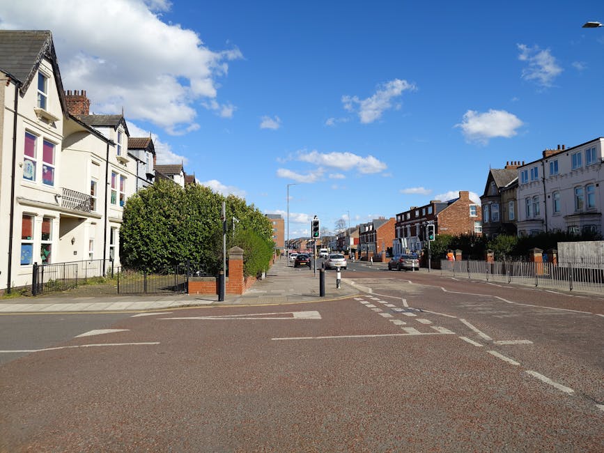 A clear view of Mottingham Lane and SE9 in daylight, showing a residential street with white and brown brick houses on both sides. The street features pedestrian crossings with white dashed lines and traffic lights, some vehicles parked along the curb and driving down the road. On the left, there is a small driveway with a black metal fence, and a large leafy green tree partly obscures some houses. The right side includes more houses with small front gardens, some with fences, and a few trees. The sky above is mostly blue with scattered clouds, and the scene is well lit, emphasizing the tidy, urban residential environment suitable for home relocation or furniture transport activities.