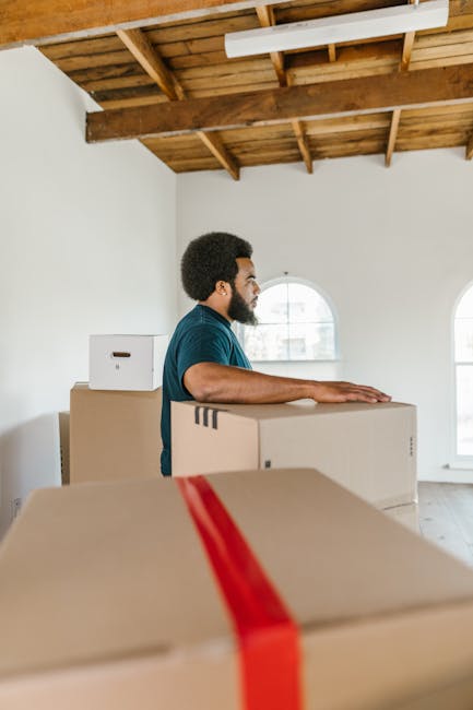 A man with dark curly hair and a beard, wearing a teal t-shirt, is standing inside a bright, spacious room with white walls and a wooden, exposed ceiling structure. He is positioned behind a stack of cardboard boxes, some of which are sealed with red packing tape, indicating they are ready for moving or transportation. The boxes vary in size and are arranged in a way that suggests he is preparing to load or carry them. Natural light is streaming in through two arched windows in the background. The room appears to be in the process of a home relocation, with minimal furniture or furnishings visible, emphasizing the packing and moving process. The setting demonstrates a professional approach to furniture transport and home removal services typical for [COMPANY_NAME], related to house removals and logistical planning.