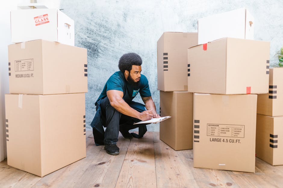 A man with dark curly hair and a beard, dressed in a blue t-shirt and black trousers, kneeling on a wooden floor inside a property, is seen writing on a clipboard. He is surrounded by several large cardboard boxes, some labeled with room contents and sizes, stacked on a bare wall with a textured surface, indicating a home relocation process. The boxes are sealed with tape, and some contain fragile items indicated by labels such as 'GLASS.' The setting suggests an ongoing packing or packing check process as part of a house removal or moving service. In the background, a green potted plant is visible on a surface near the doorway. The lighting appears natural, highlighting the details of the packing materials and the man's focused activity, characteristic of professional removals and furniture transport activities by Man with Van Mottingham.