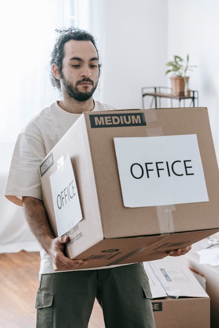 A man with dark, curly hair and a beard is indoors, carefully holding a large cardboard box labeled 'OFFICE' with a 'MEDIUM' size indication, preparing for a home relocation or office move. The box is sealed with packing tape, and the man is gripping it securely with both hands, standing on a wooden floor. In the background, there are additional boxes stacked on the floor, some also labeled, as well as a window letting in natural light and a small table with a potted plant on it. The scene captures the packing and loading process typical of house removals or business relocations, which could be managed by professional services such as Man with Van Mottingham. This setting highlights the careful handling and organization involved in furniture transport, packing, and moving logistics relevant to small business relocations in the Mottingham area.