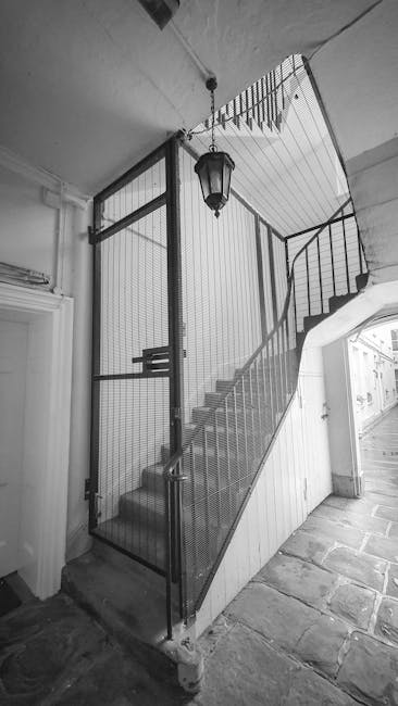 The image shows a narrow interior staircase inside a property, with metal framing and mesh panels forming a secure staircase enclosure. The staircase features a metal handrail on the right side, leading upward with visible steps made of wood or a similar material, and is positioned adjacent to an open door with a glass window. The ceiling above has a hanging lantern-style light fixture, and the floor at the base of the stairs is made of stone or tile. The scene appears to be part of a house removal process, where careful packing and moving strategies are required for confined staircases, as managed by Man with Van Mottingham, who specializes in residential furniture transport and home relocation services. The enclosed staircase is designed to facilitate efficient and safe furniture transport during a packing and moving operation, with the clear layout and existing protective materials supporting effective logistics within the property.