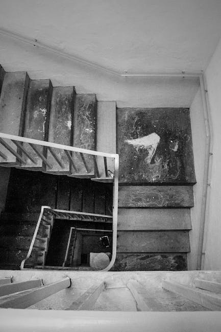 The image shows a narrow interior staircase inside a property, with metal framing and mesh panels forming a secure staircase enclosure. The staircase features a metal handrail on the right side, leading upward with visible steps made of wood or a similar material, and is positioned adjacent to an open door with a glass window. The ceiling above has a hanging lantern-style light fixture, and the floor at the base of the stairs is made of stone or tile. The scene appears to be part of a house removal process, where careful packing and moving strategies are required for confined staircases, as managed by Man with Van Mottingham, who specializes in residential furniture transport and home relocation services. The enclosed staircase is designed to facilitate efficient and safe furniture transport during a packing and moving operation, with the clear layout and existing protective materials supporting effective logistics within the property.